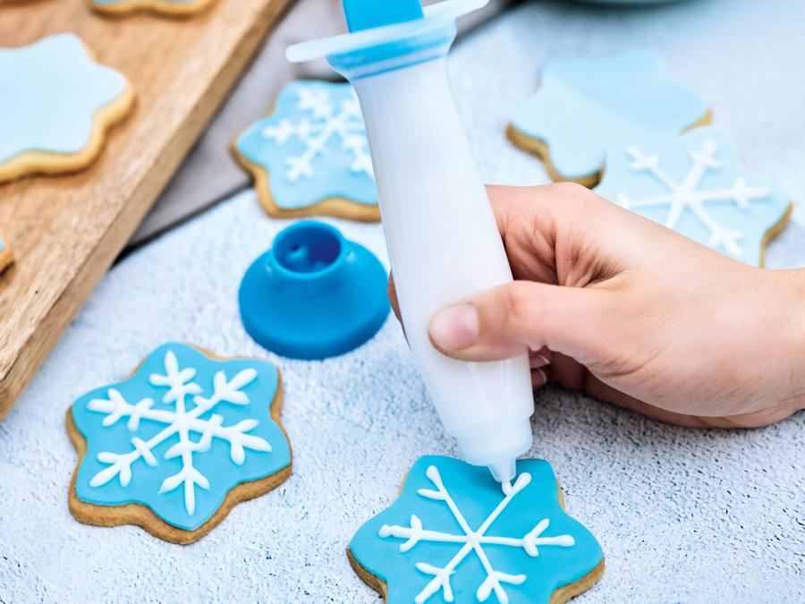 white and blue deco pen filled with white icing being held in a womans hand and used to decorate christmas cookies with blue and white icing in the form of a snowflake