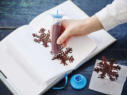 woman holding the tupperware deco pen with chocolate frosting and using it to make chocolate snowflake desserts on a napkin placed in a book